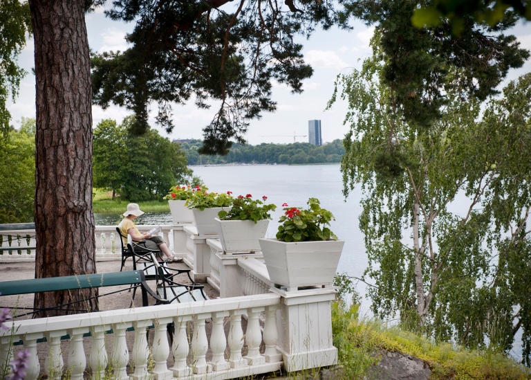 Italienska terrassen med vit balustrad. En kvinna sitter på en bänk och läser. Vy över Brunnsviken.